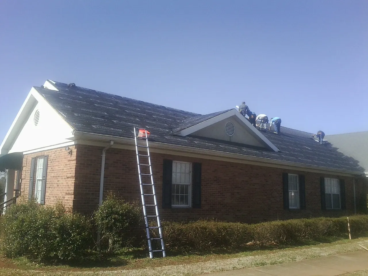 Crew tearing shingles off a brick ranch roof with a ladder set — Metal Roof Repair work by our crew in Arnoldsburg