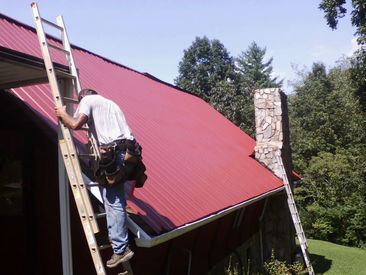 Roofer on a ladder beside a red metal roof and stone chimney — Soffit & Fascia Repair by Roofing Brothers in Arnoldsburg
